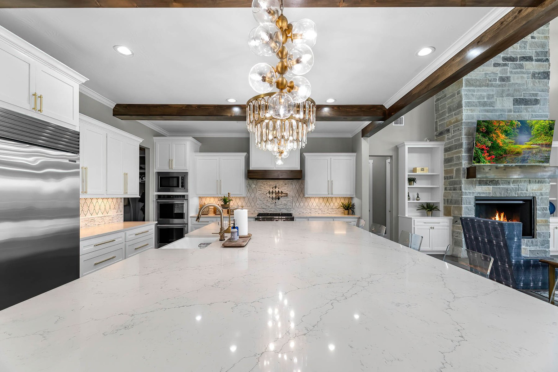 White kitchen with a quartz island, globe chandelier, wood ceiling beams and a stone fireplace